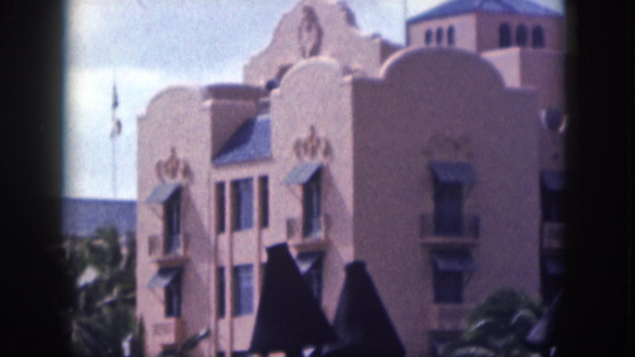 Spanish Colonial Revival style facade with shaped parapets, awnings, palms, and foreground lamps.