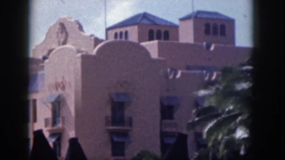 1959 Hawaii 8mm view of a pale stucco facade with arched parapet forms, tower windows, palms, and dark lamps.