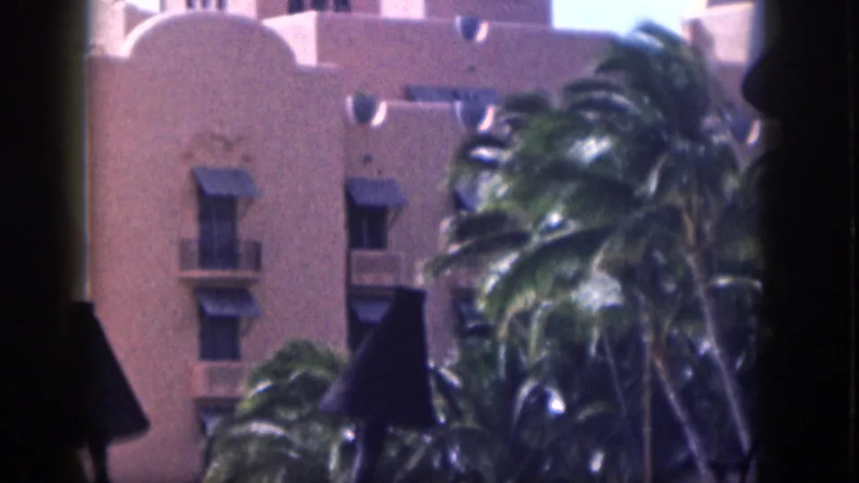 1959 Hawaii frame with wind-bent palms and dark conical lamps in front of a pale stucco building facade.