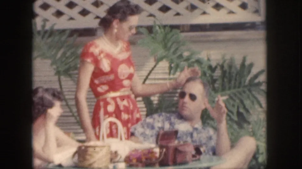 1959 Hawaii patio scene with a woman in a red floral dress standing beside a reclining man in sunglasses and another seated person.