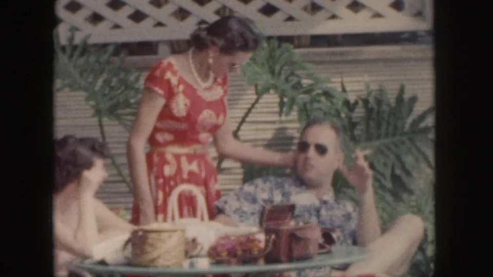 1959 Hawaii home-movie frame of friends on a patio, including a woman in a red floral dress and a man reclining with sunglasses.