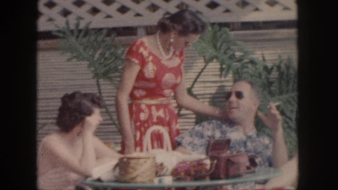 1959 Hawaii home-movie frame showing adults gathered at a patio table, with a woman in a red patterned dress beside a reclining man in sunglasses.