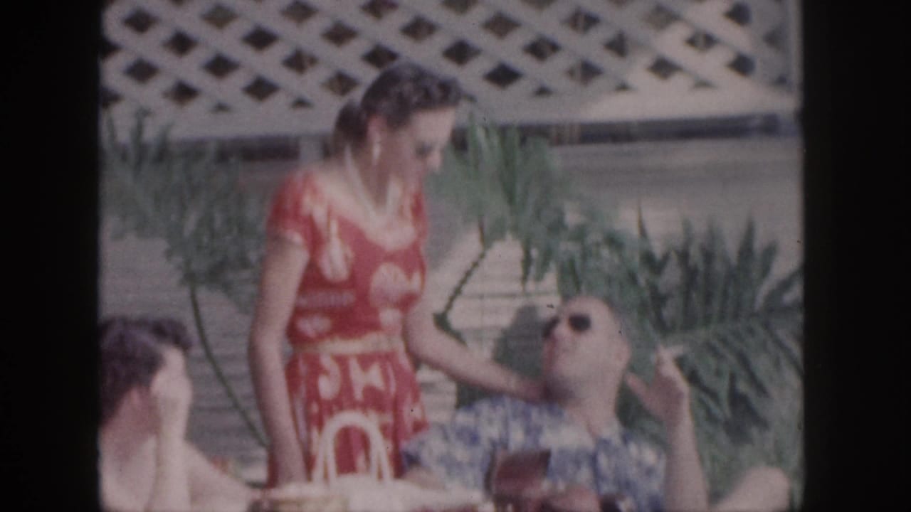 Woman in a red dress stands beside a reclining man in sunglasses while another person sits at left.