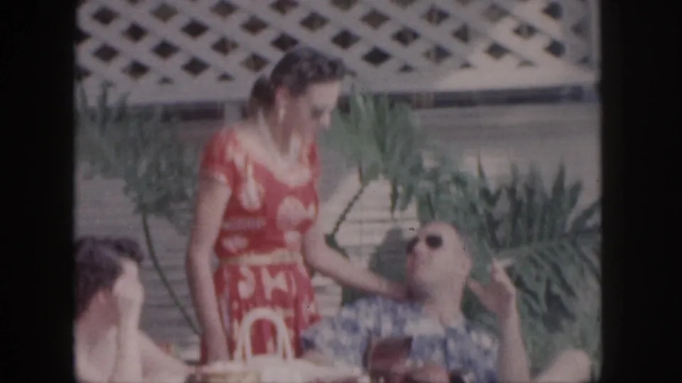 1959 Hawaii vacation frame with a woman in a red patterned dress standing near a reclining man in a Hawaiian shirt and sunglasses.