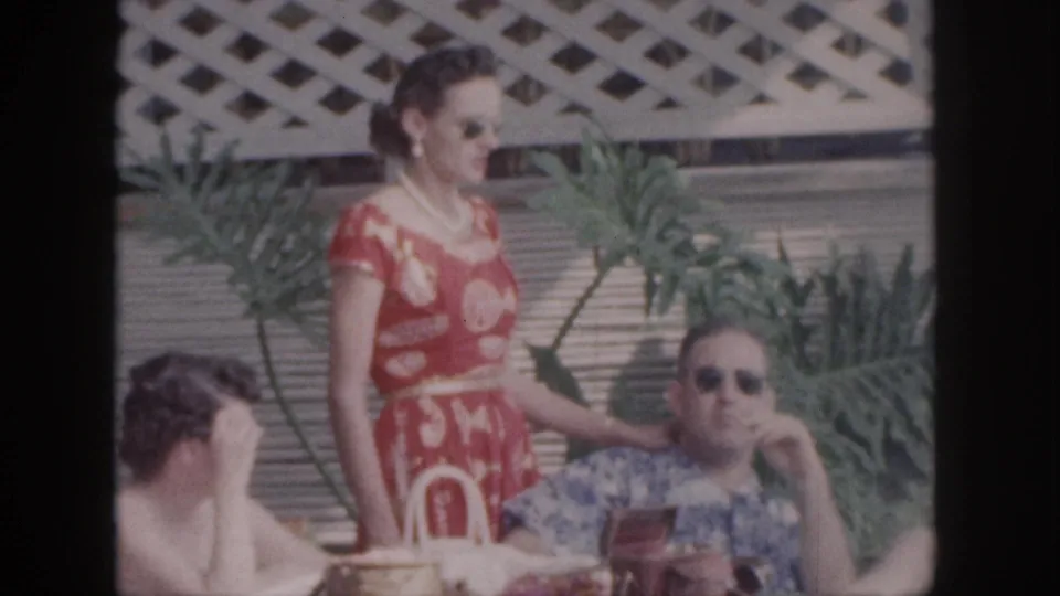 1959 Hawaii home-movie frame showing a patio gathering with tropical plants, a red dress, sunglasses, and food on the table.