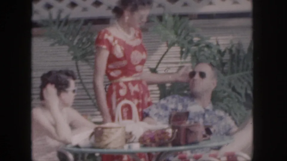 1959 Hawaii vacation frame with three adults on a patio, including a woman in a red dress and a man in sunglasses by a table.