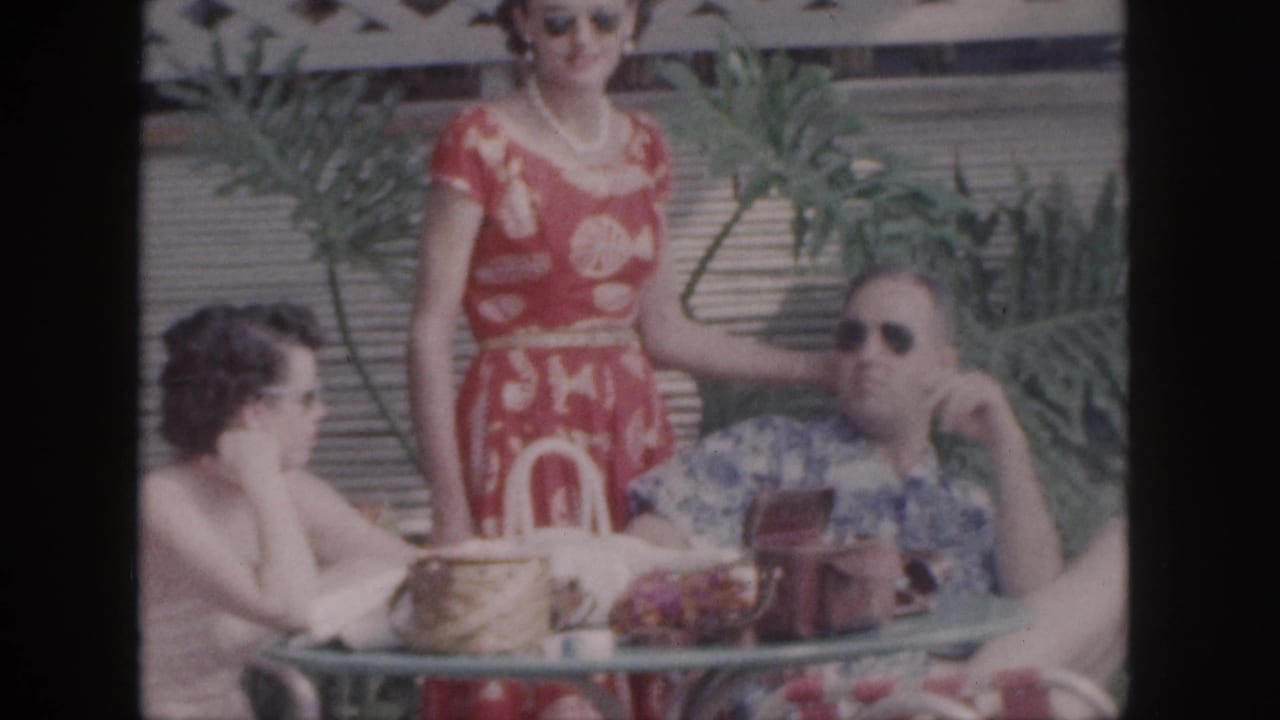 Three adults at an outdoor table, with a woman in a red dress standing beside a seated man.