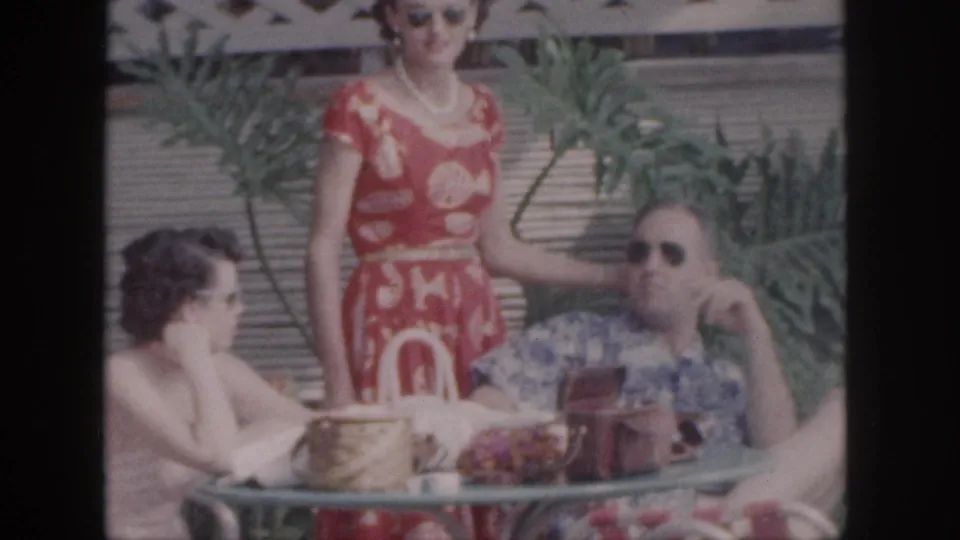 1959 Hawaii home-movie frame of a woman in a red patterned dress standing by a seated man in sunglasses while another woman sits at the table.