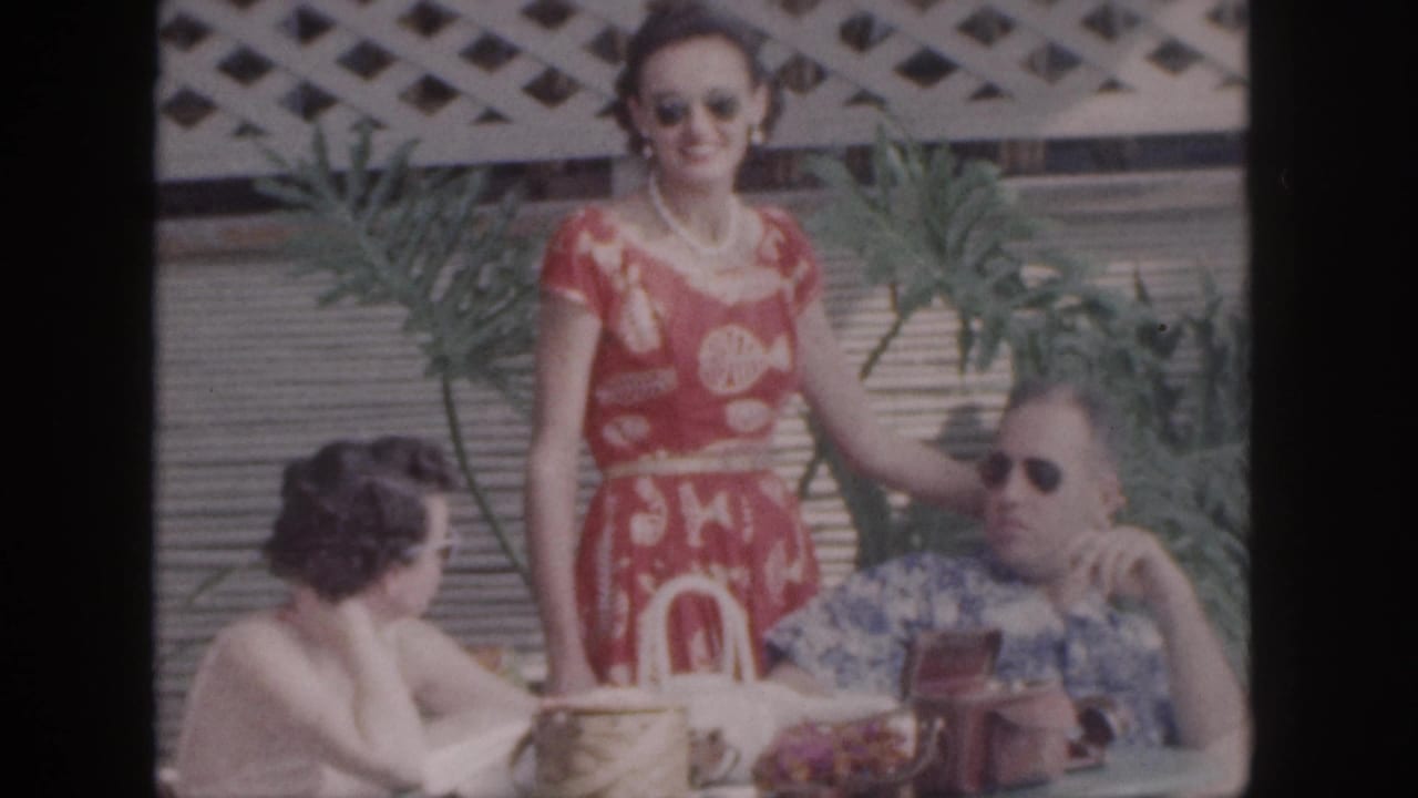 Woman in red patterned dress smiles beside a seated man while another woman sits at the table.
