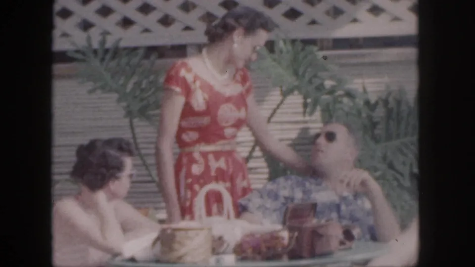 1959 Hawaii home-movie frame of the standing woman in a red dress leaning slightly toward the seated man, with a second woman nearby.