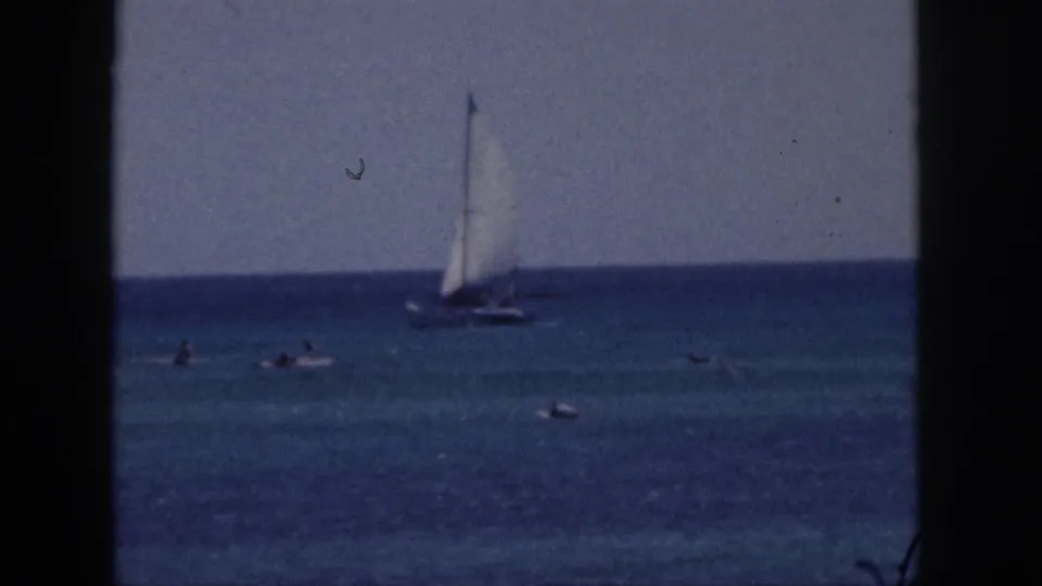 1959 Hawaii home-movie cutaway of a sailboat on blue ocean water, with swimmers scattered in the foreground under a clear sky.
