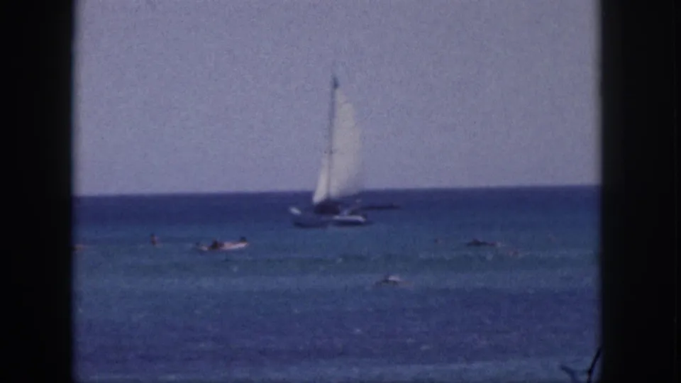 1959 Hawaii 8mm frame showing a small sailboat beyond distant swimmers in calm ocean water.