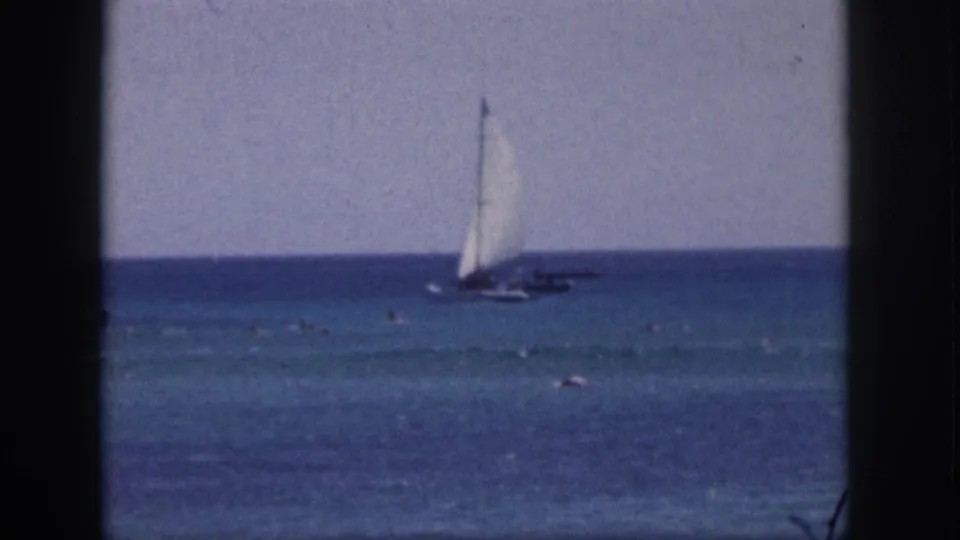 1959 Hawaii home-movie frame with a white-sailed boat offshore and small swimmers visible in the water.