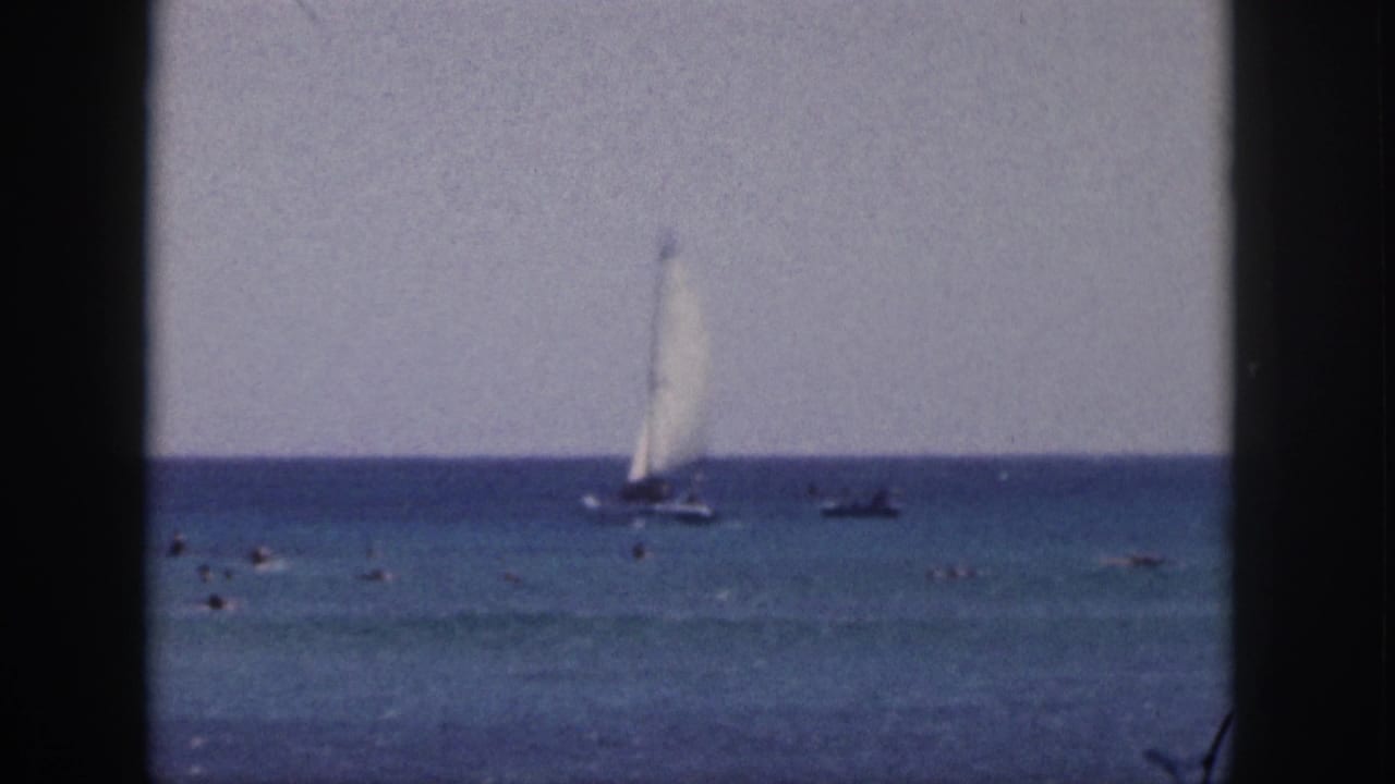1959 Hawaii home-movie frame showing a sailboat offshore, scattered swimmers, pale sky, and dark film-frame edges.