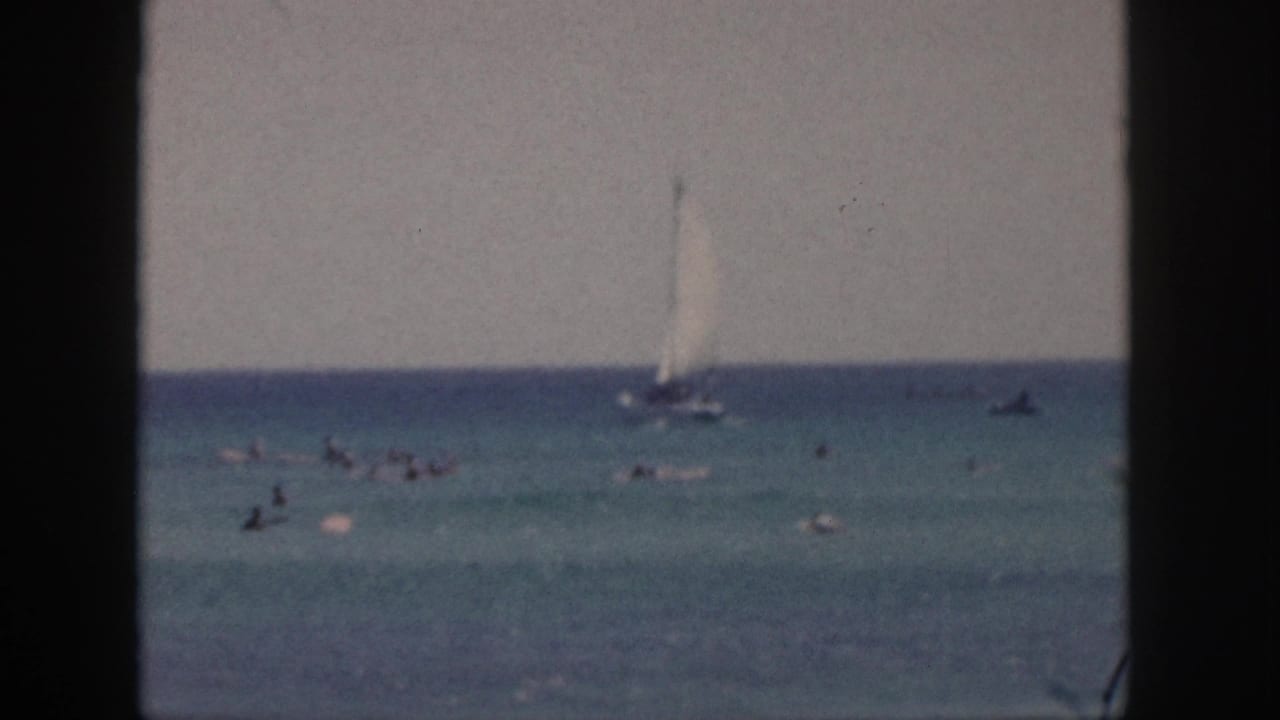 1959 Hawaii beach footage frame with a white sailboat, distant swimmers, small boats, and soft color 8mm texture.