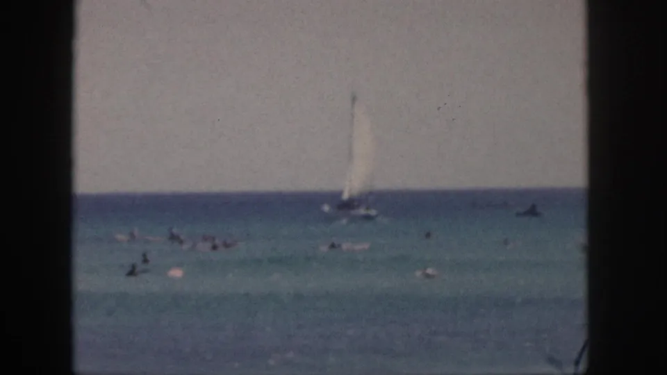 1959 Hawaii beach footage frame with a white sailboat, distant swimmers, small boats, and soft color 8mm texture.