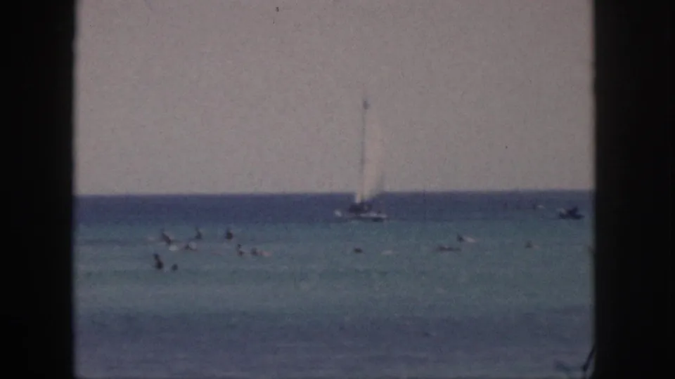 1959 Hawaii beach scene with distant swimmers, a white sailboat offshore, small boats, and grainy 8mm color.
