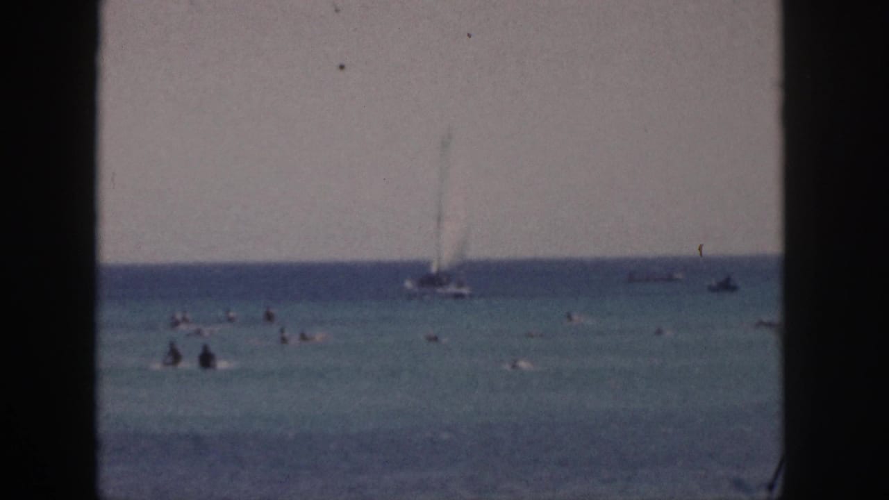 1959 Hawaii 8mm frame showing swimmers in clear ocean water, a sailboat on the horizon, and dark foreground framing from shore.