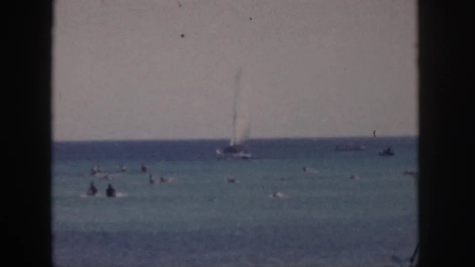 1959 Hawaii 8mm frame showing swimmers in clear ocean water, a sailboat on the horizon, and dark foreground framing from shore.