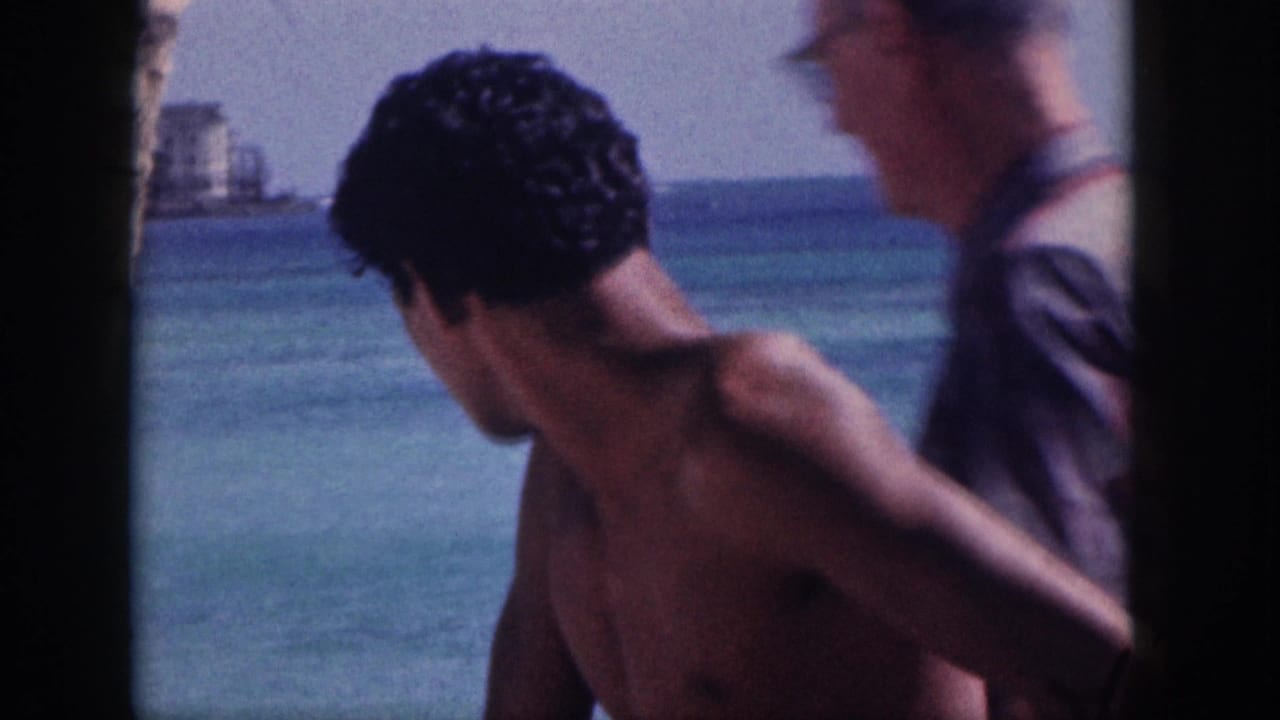 Shirtless man near the water with another adult partly visible beside him.