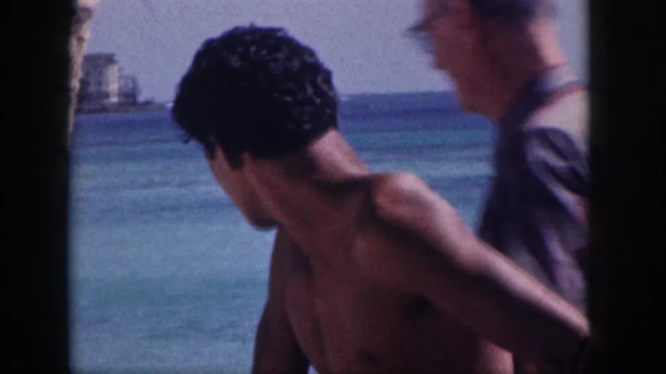 1959 Hawaii beach frame with a shirtless man in the foreground, another adult at the edge, turquoise water, and distant shoreline buildings.