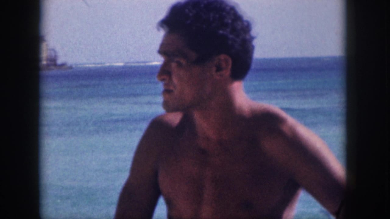 1959 Hawaii 8mm frame showing a shirtless man in profile near the beach, with calm ocean water and distant coastal buildings behind him.