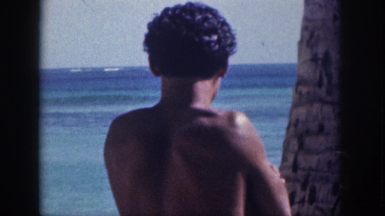 1959 Hawaii home-movie frame of a curly-haired shirtless man from behind, standing near a palm trunk and ocean water.