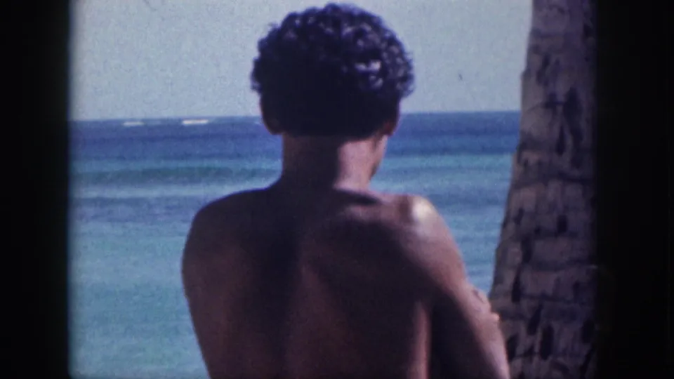 1959 Hawaii home-movie frame of a curly-haired shirtless man from behind, standing near a palm trunk and ocean water.