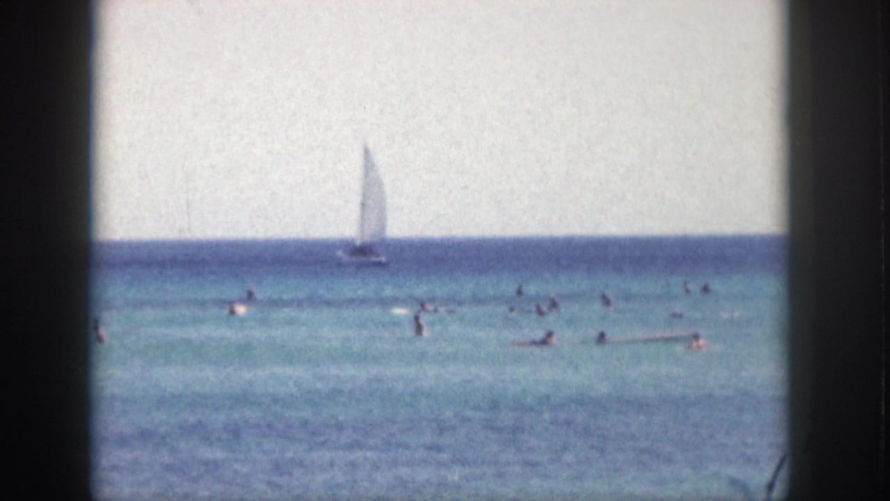1959 Hawaii coastal frame with swimmers spread across turquoise water and a small sailboat in the distance.