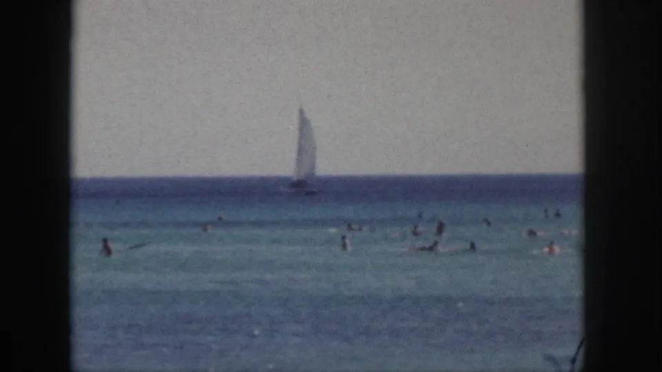 1959 Hawaii home-movie frame showing distant swimmers in blue-green ocean water and a sailboat near the horizon.