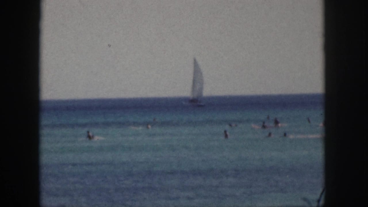 1959 Hawaii beach footage frame with small swimmers in the water and a sailboat moving along the far horizon.