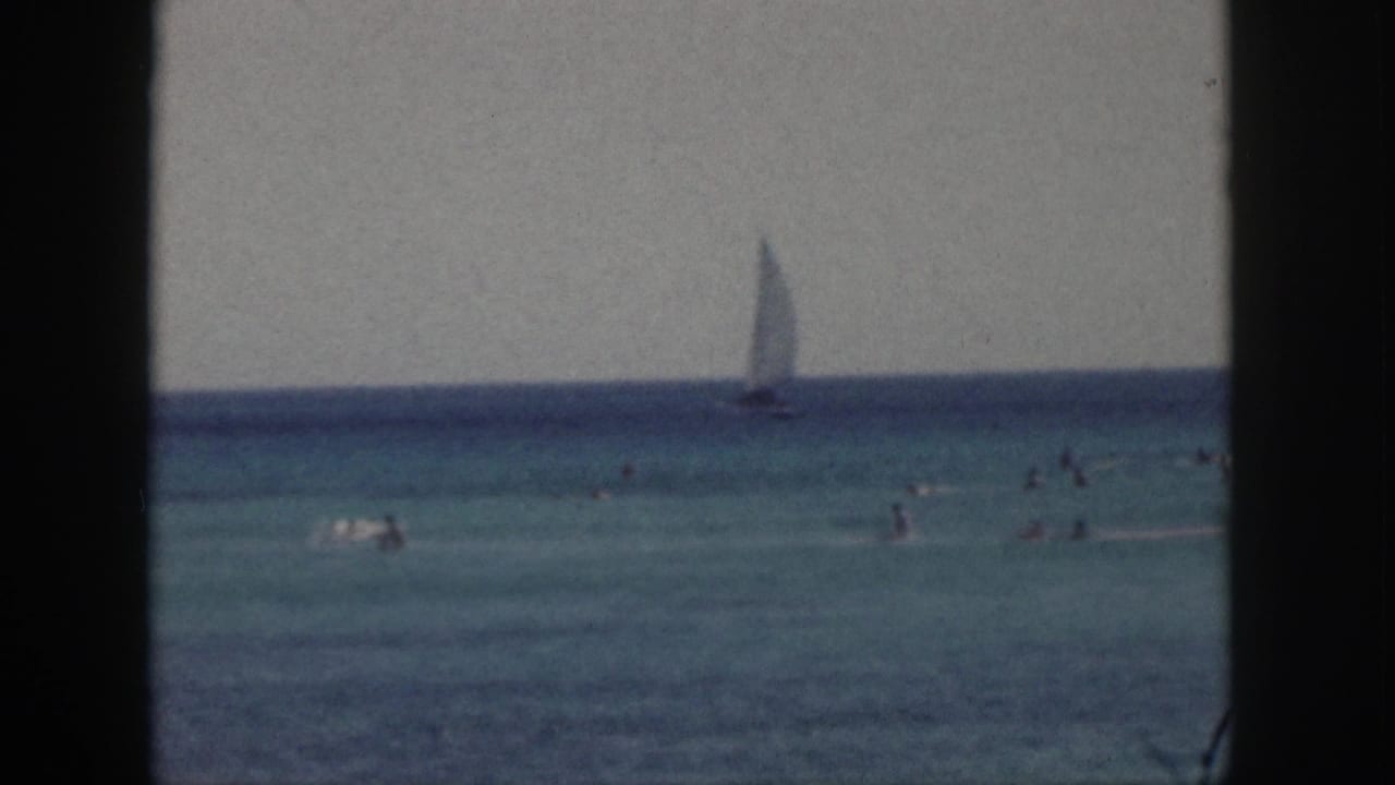 1959 Hawaii 8mm frame showing beachgoers swimming in blue-green water with a sailboat offshore.