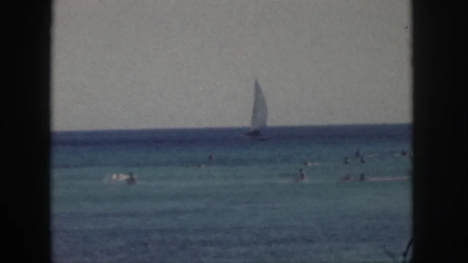 1959 Hawaii 8mm frame showing beachgoers swimming in blue-green water with a sailboat offshore.