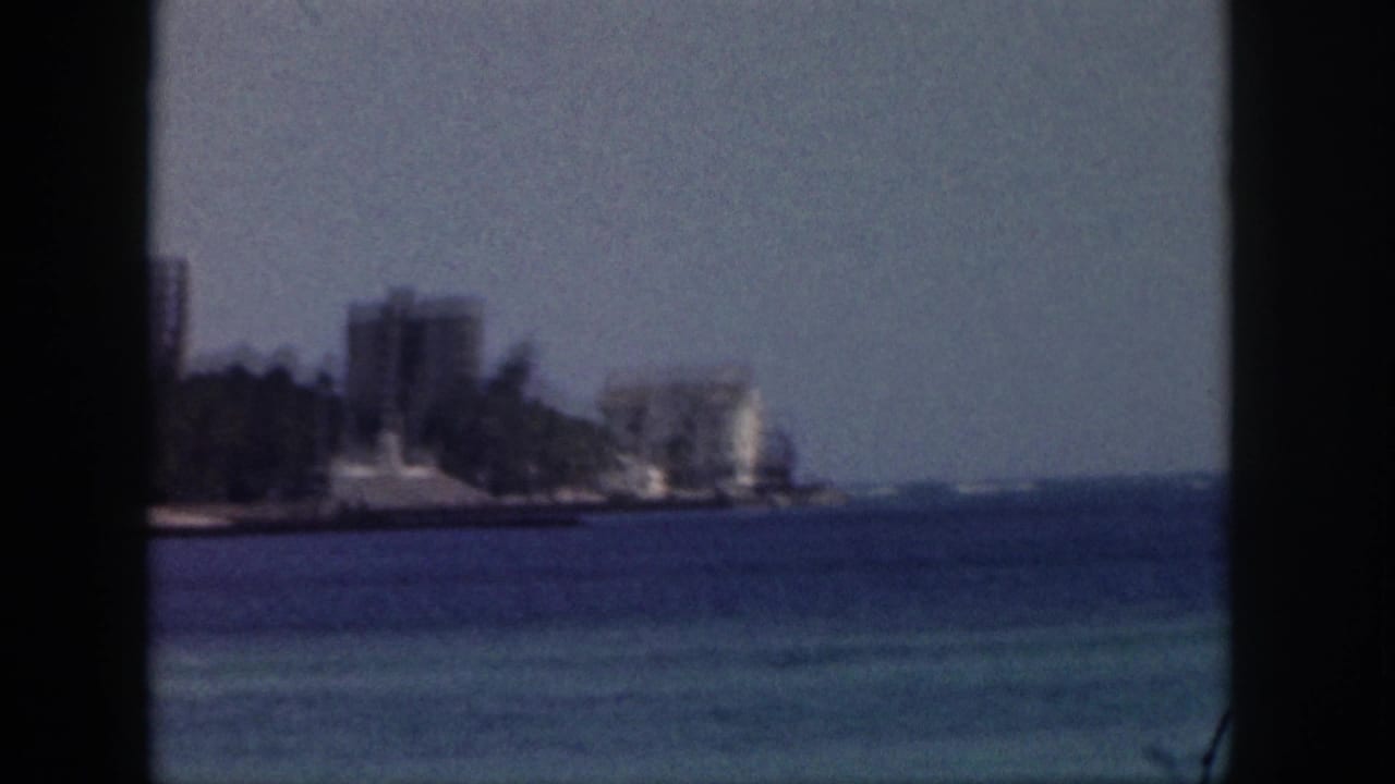 1959 Hawaii coastal view with distant mid-century buildings, palm trees, blue water, and dark vertical window framing.