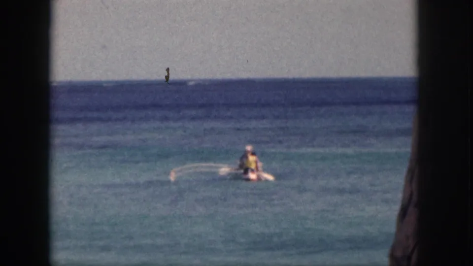 1959 Hawaii home-movie frame of a distant paddler crossing turquoise water below a dark blue horizon.