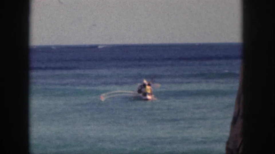 Distant Hawaii paddling scene with a small craft, visible wake arcs, blue water, and 8mm home-movie borders.