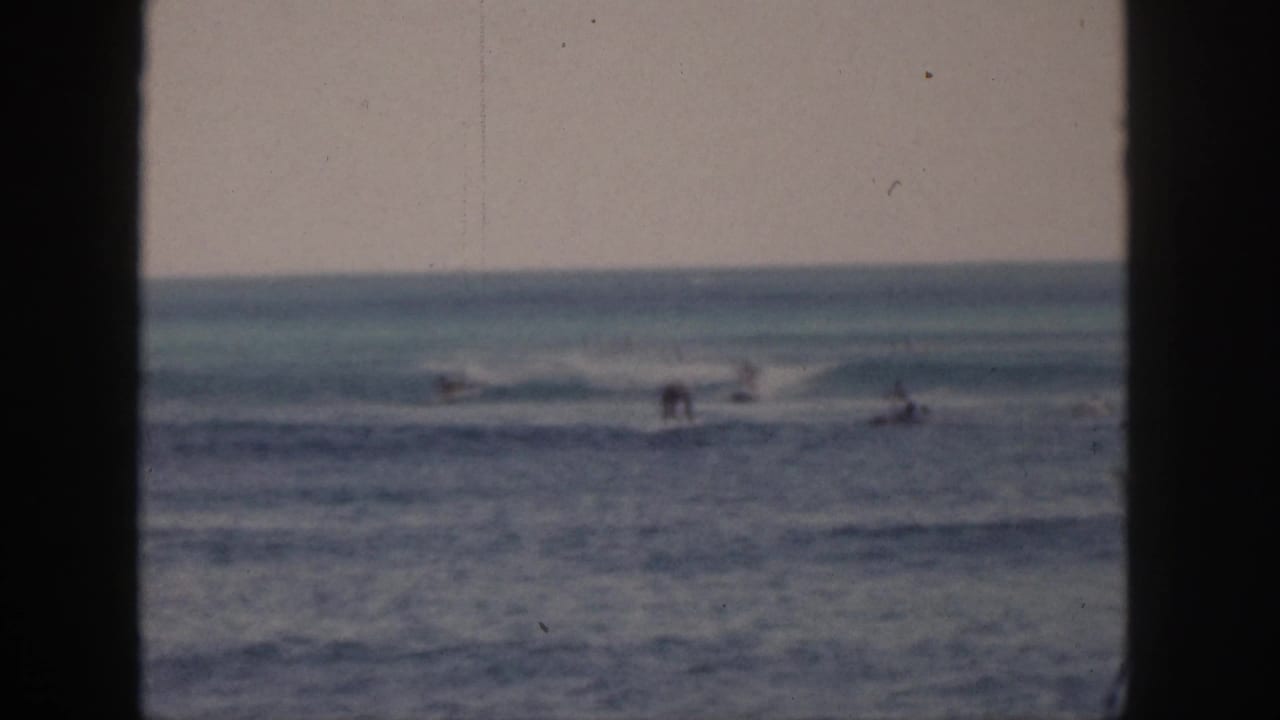 1959 Hawaii 8mm frame showing distant surfers and swimmers beyond a dark window or doorway edge, with breaking waves and soft color fade.