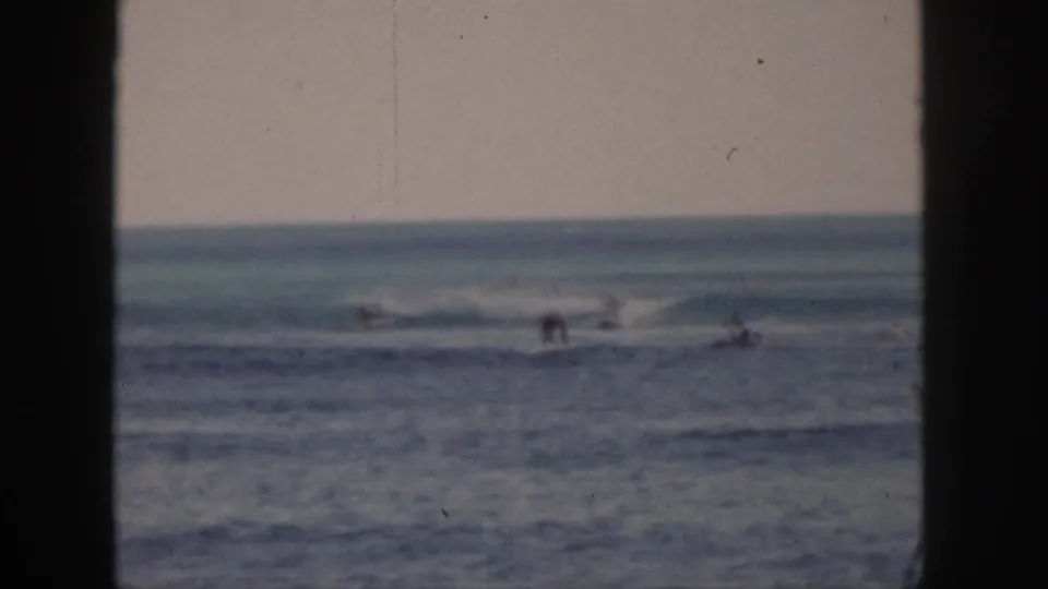 1959 Hawaii 8mm frame showing distant surfers and swimmers beyond a dark window or doorway edge, with breaking waves and soft color fade.