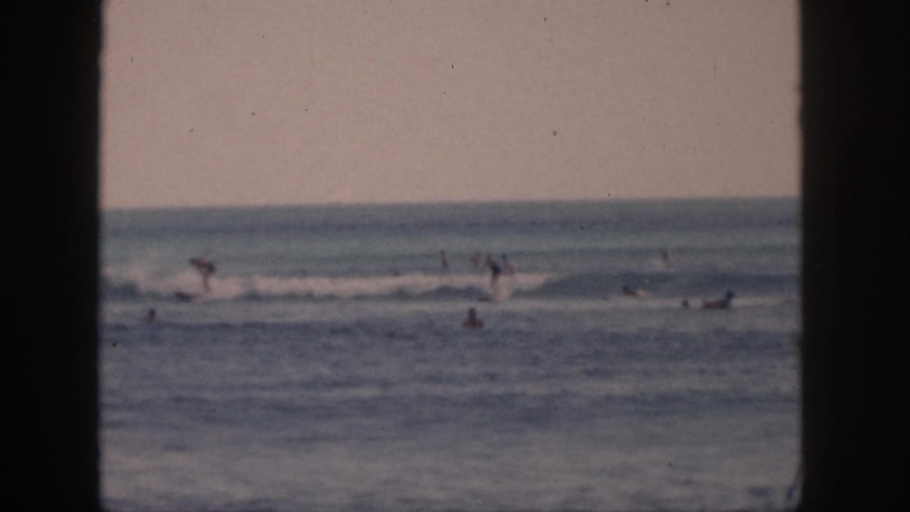 Distant surfers and swimmers move through Hawaii ocean waves in a 1959 8mm home-movie view framed by dark vertical edges.