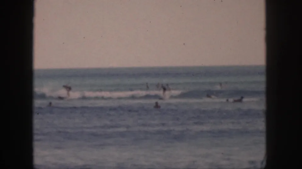 Distant surfers and swimmers move through Hawaii ocean waves in a 1959 8mm home-movie view framed by dark vertical edges.