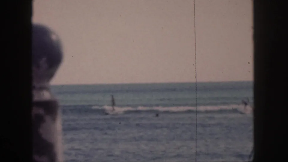 A 1959 Hawaii beach view shows distant swimmers in the surf, with a partial foreground person at the left edge and visible 8mm film wear.