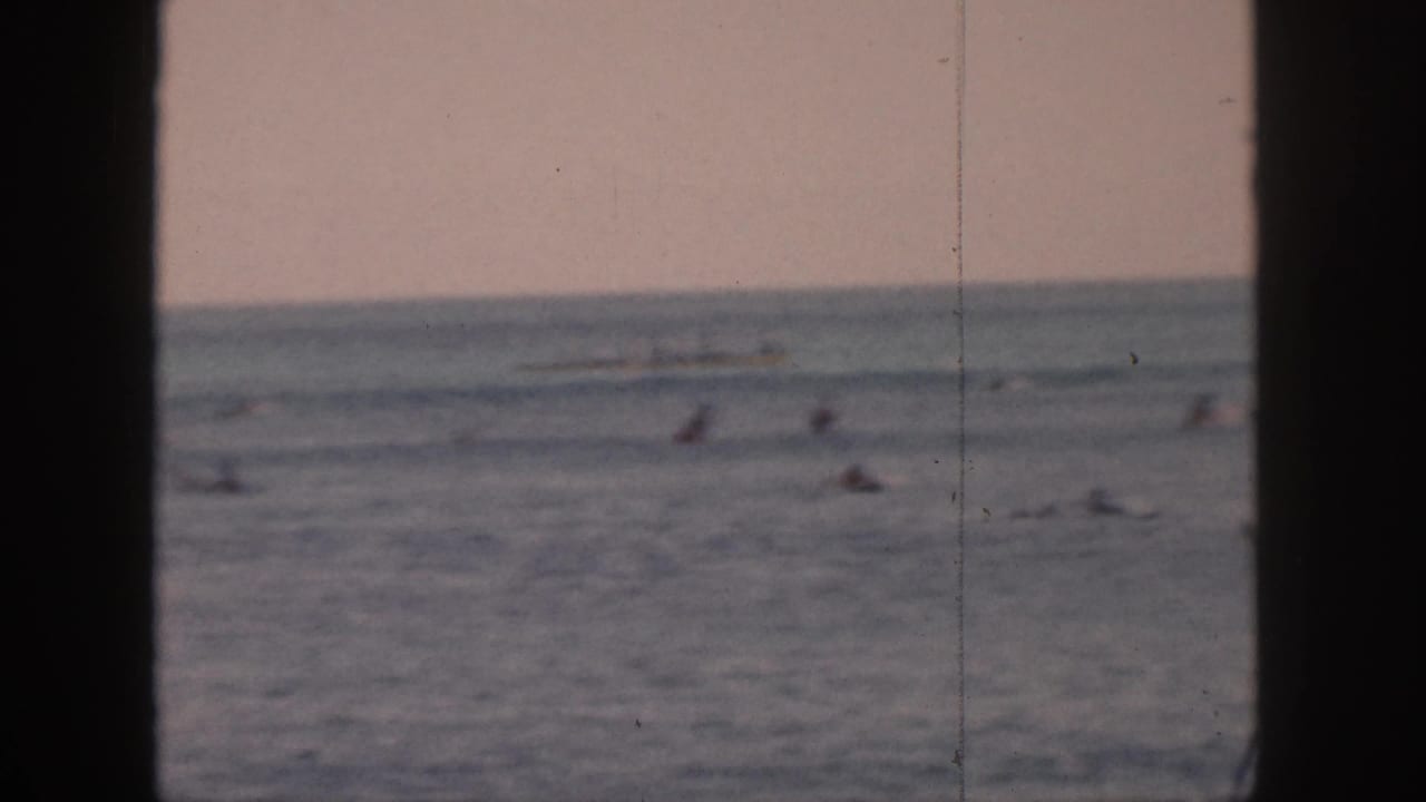 1959 Hawaii 8mm frame showing distant surfers scattered offshore, viewed through dark vertical framing with a soft horizon.