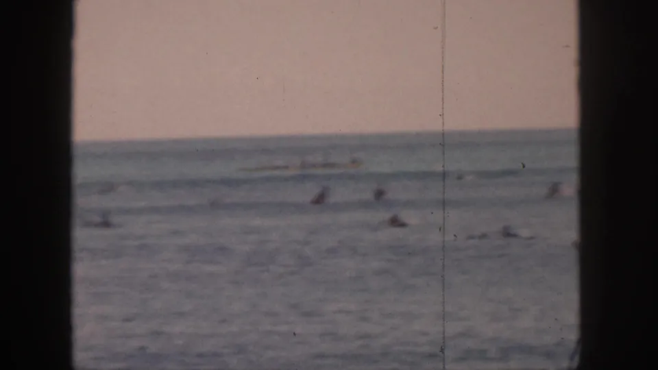 1959 Hawaii 8mm frame showing distant surfers scattered offshore, viewed through dark vertical framing with a soft horizon.