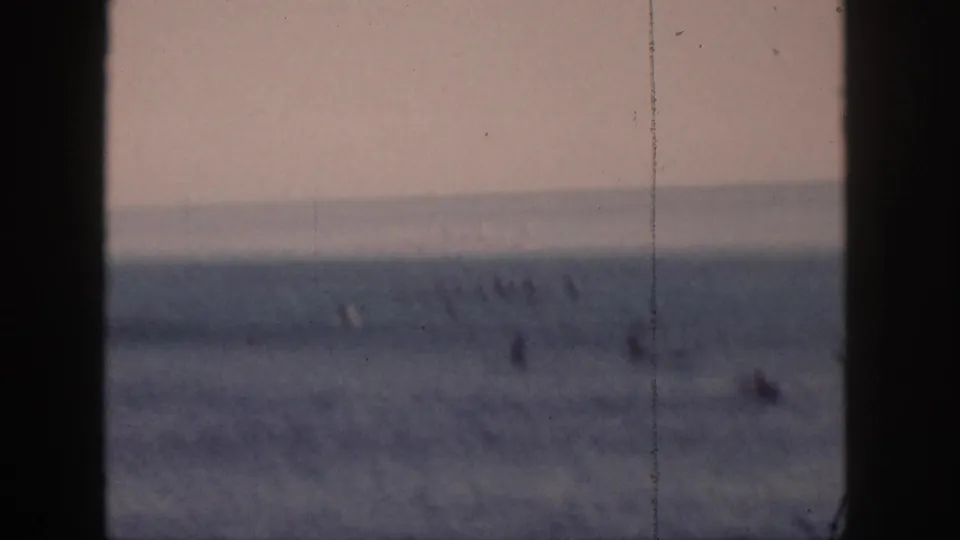 Distant 1959 Hawaii surf scene with small figures on boards across the water and dark foreground bars dividing the view.