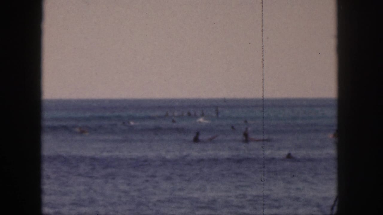1959 Hawaii home-movie frame with multiple surfers waiting on boards in a muted blue ocean under a pale sky.