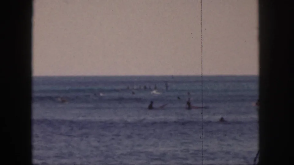 1959 Hawaii home-movie frame with multiple surfers waiting on boards in a muted blue ocean under a pale sky.