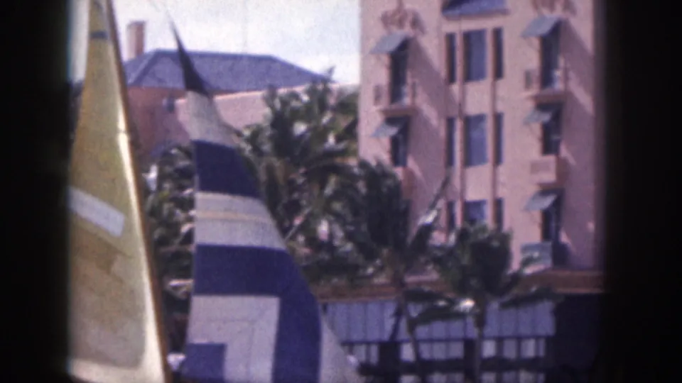 1959 Hawaii 8mm frame with yellow and striped sailboat sails, palm trees, and a pink hotel facade in sunlight.