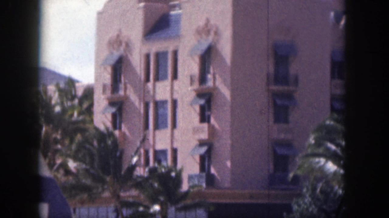 Pink hotel facade with palm trees and dark awnings.