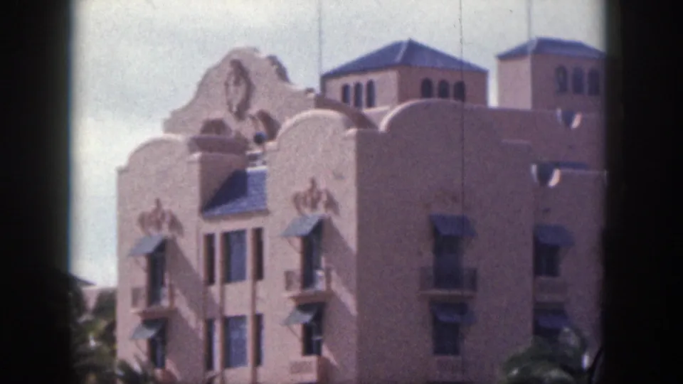 1959 Hawaii 8mm frame of an ornate pink hotel exterior with curved roofline details, windows, awnings, and palms.