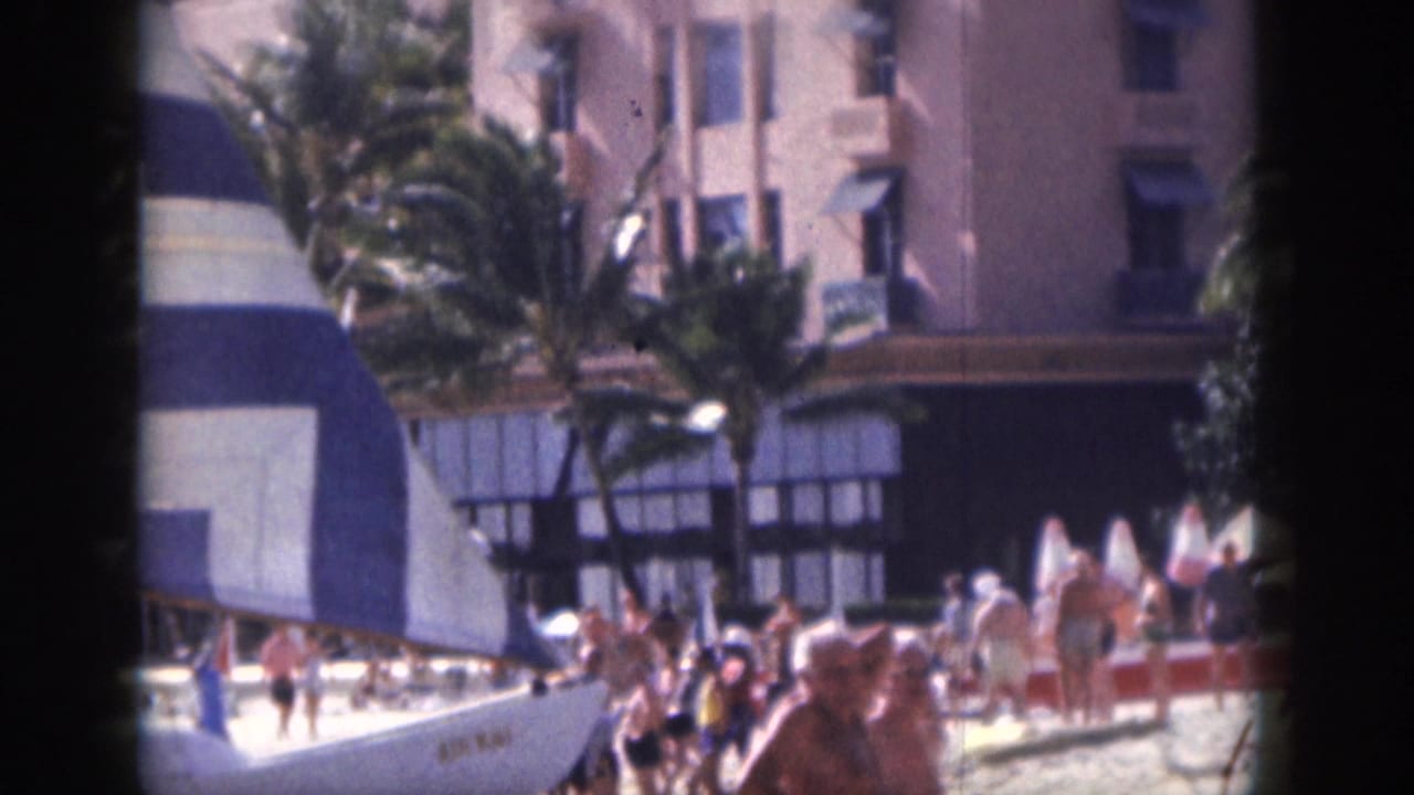 1959 Hawaii beach scene with people gathered near umbrellas, a striped sail, palm trees, and the pink building behind them.